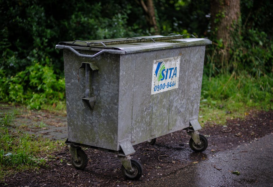 A weathered metal waste bin with a blue and white SITA logo and contact number, situated on a damp, uneven outdoor surface near a garden boundary. The bin has a closed lid with a handle, and tilted wh