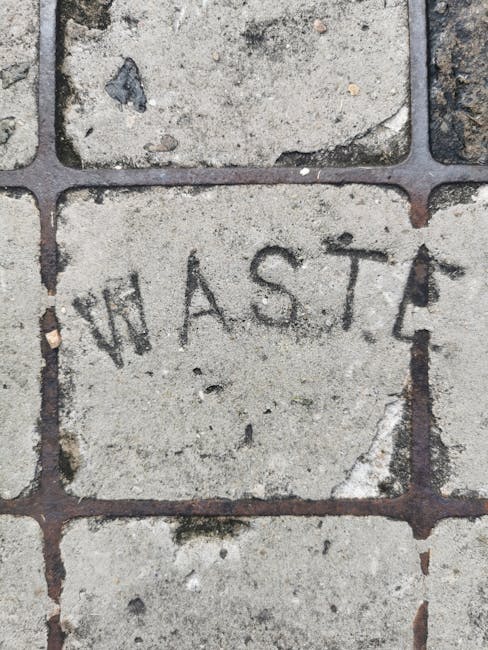 A weathered metal waste bin with a blue and white SITA logo and contact number, situated on a damp, uneven outdoor surface near a garden boundary. The bin has a closed lid with a handle, and tilted wh