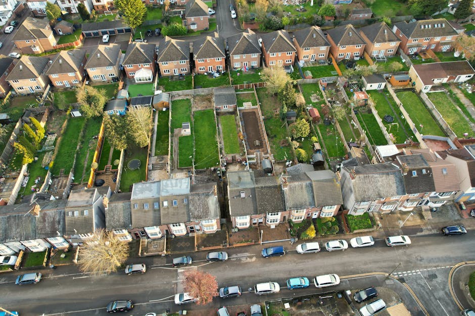 An aerial view of a residential street with a row of terraced houses featuring tiled roofs and small front gardens. Behind the houses, there are numerous backyards and garden spaces, some with lush gr